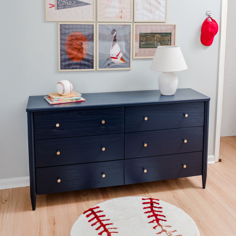 Navy blue six-drawer dresser in kids' sports-themed bedroom with white table lamp, baseball rug, stacked children's books and framed sports wall art
