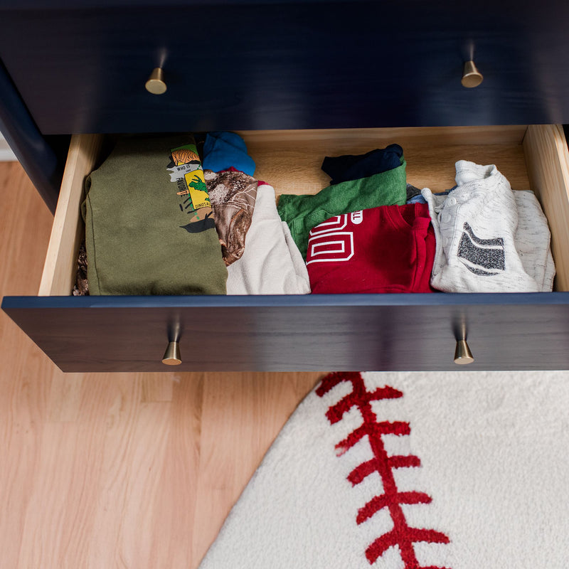 Open navy dresser drawer filled with neatly folded children's shirts and pajamas beside a baseball-stitched round rug on light hardwood floor