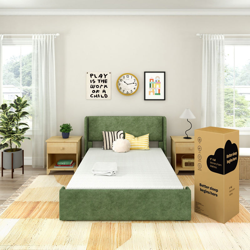 Green upholstered bed with boxed memory foam mattress in a bright modern bedroom with wooden nightstands, lamp, houseplant, decorative pillows and children's wall art