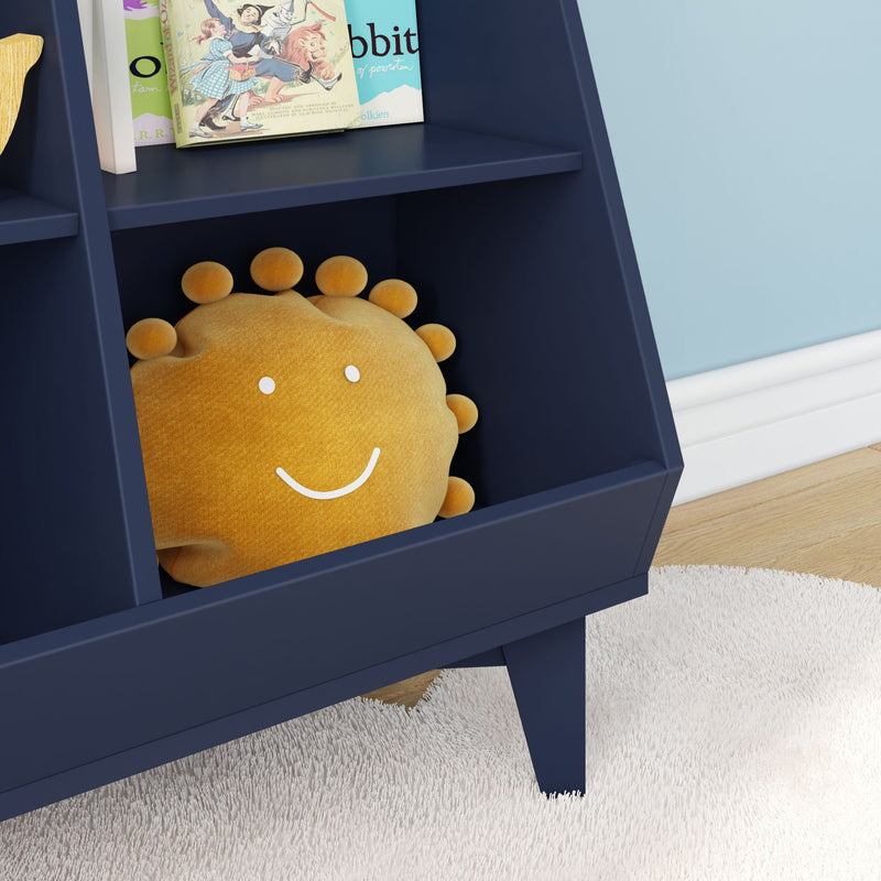 Navy blue kids bookshelf in a modern nursery holding a smiling yellow sun plush pillow and illustrated children's books, set against a light blue wall with white shag rug and hardwood floor.