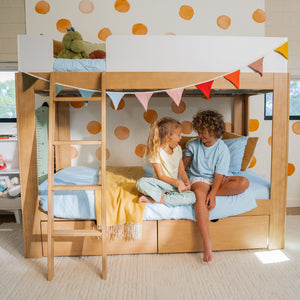 Children laughing on a wooden bunk bed with ladder, pastel bedding and bunting, polka-dot wallpaper and a stuffed dinosaur