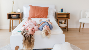 Two people lying upside down on a white twin mattress in a bright modern bedroom with wooden nightstands and a terracotta pillow