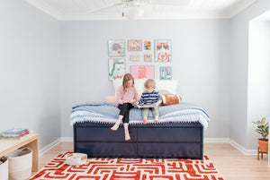 Two children reading on a navy storage bed in a bright modern kids' bedroom with pastel animal wall art, blue quilted bedding, and a red geometric rug
