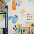 Children's bedroom decor: navy bunk bed, colorful abstract wall mural, wooden cubby shelf with clock, books, toy dinosaur and canvas sign reading "COOL KIDS LIVE HERE