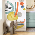 Modern nursery with colorful abstract watercolor wall mural, gray elephant toy, mint green dresser with round mirror and changing pad — stylish kids room decor