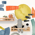 Modern nursery wall decor with colorful watercolor decals, light wood floating shelves displaying a knitted gray plush toy, Brown Bear board book, small potted plant and white baby monitor camera