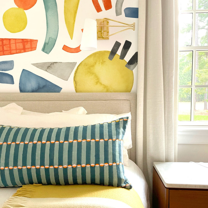 Modern bedroom with colorful watercolor mural accent wall, beige upholstered headboard, teal striped lumbar pillow, cream curtains and wooden bedside table by window