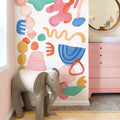 Colorful abstract watercolor nursery wall mural with pink changing dresser and elephant stool, modern kids' room decor