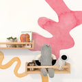 Nursery wall with pink watercolor mural and light wood floating shelves displaying the book "Brown Bear, Brown Bear, What Do You See?", a small succulent in a white planter, wooden toy car, gray knitted doll and white baby monitor — modern kids room decor and wooden toy shelf styling