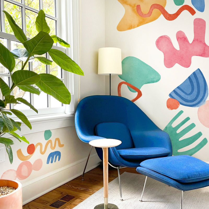 Cozy modern reading nook with blue lounge chair and ottoman, colorful abstract wall mural, fiddle leaf fig plant by window, floor lamp and small round side table