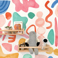 Colorful nursery wall mural with watercolor abstract shapes, wooden floating shelves, stuffed toy, baby monitor and Brown Bear board book