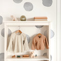 Neutral baby nursery wardrobe with hanging beige striped dress and peach cardigan, wooden toys and plush bear on white shelf, minimalist polka-dot wall decor