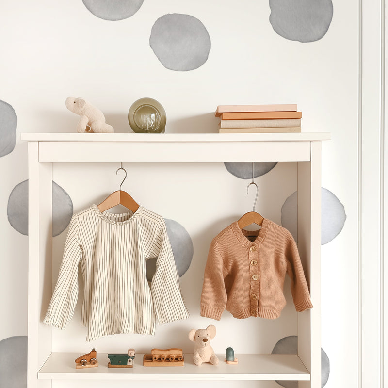 Neutral baby nursery wardrobe with hanging beige striped dress and peach cardigan, wooden toys and plush bear on white shelf, minimalist polka-dot wall decor