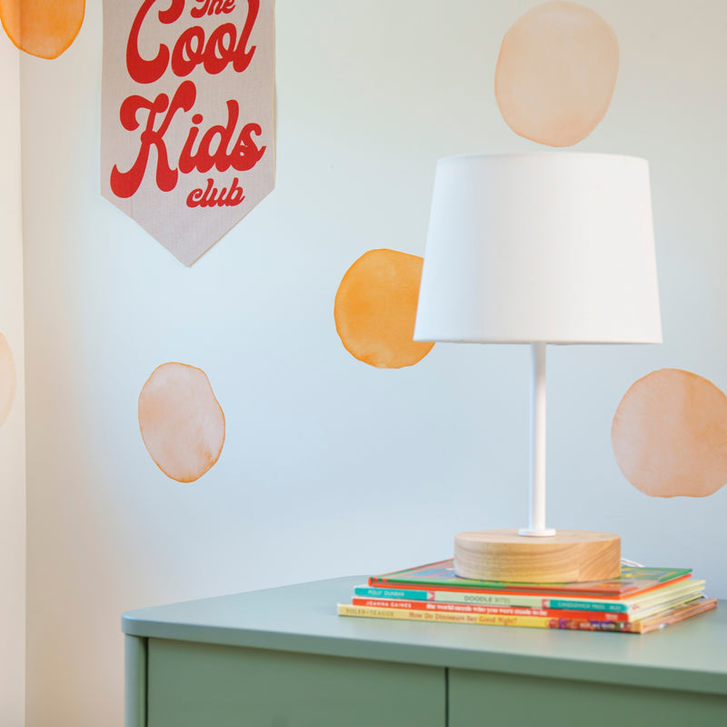 Kids room decor: white table lamp on a mint-green dresser with stacked children's books, orange watercolor circle wall decals and a "The Cool Kids Club" fabric banner