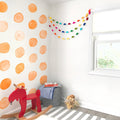 Bright modern nursery corner with orange watercolor polka dot accent wall, rainbow paper garland, red wooden rocking horse, gray storage basket with striped blanket and wire basket of wooden toys by a sunny window on a striped rug