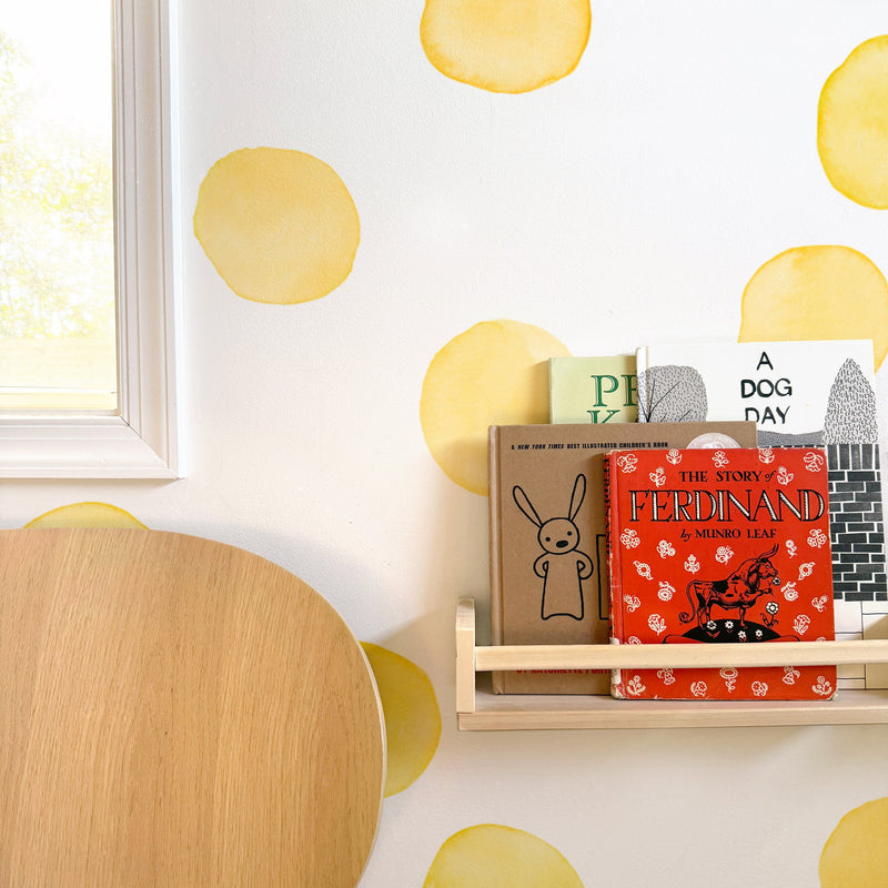 Children's books on a wooden nursery bookshelf with a red "Ferdinand" cover, round yellow polka-dot wall decals, light oak table and window light