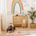 Bright children's nursery with rainbow wall mural, wooden three-drawer dresser, vintage toy car, plush bear, framed prints, and potted palm plant