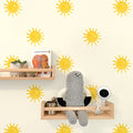 Nursery decor with yellow sun wall decals, light wood floating shelves holding a potted plant, wooden toy car, "Brown Bear" board book, knitted gray doll and white baby monitor.