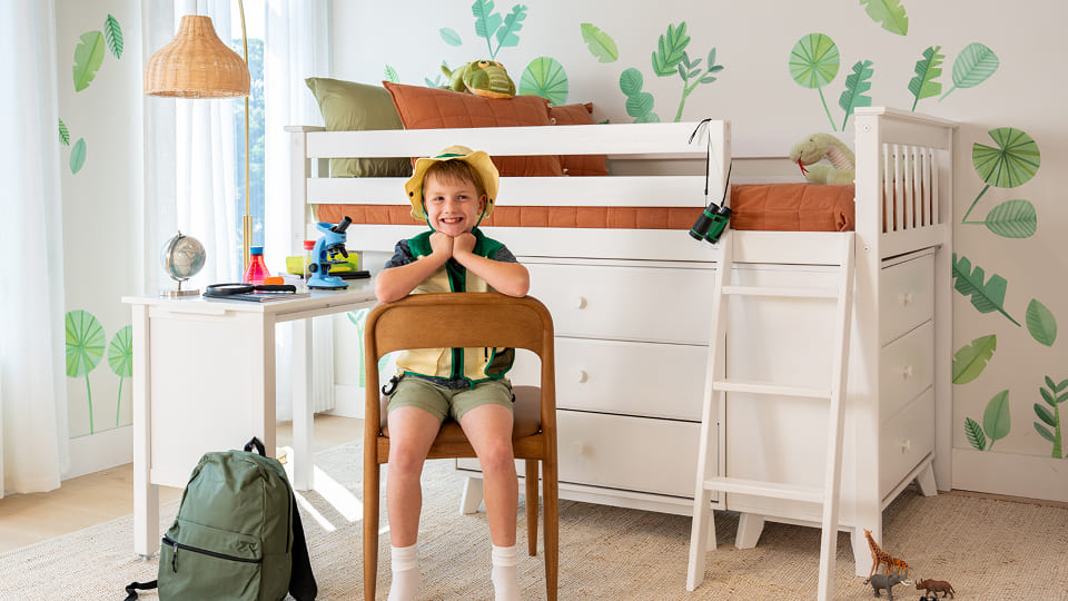 Child's bedroom with white loft bed and storage drawers, ladder and study desk with microscope and globe, green backpack, nature-themed leaf wall decals and smiling child wearing an explorer hat.