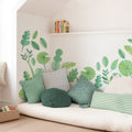 Cozy reading nook with green botanical leaf wall decals, textured green and striped throw pillows on a cream floor mattress, wicker book basket and minimalist white shelf
