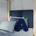 Navy blue letter "A" accent pillow on striped blue-and-white bedding with pale blue pillows and a dark upholstered headboard in a light wood canopy twin bed — modern coastal kids' bedroom decor