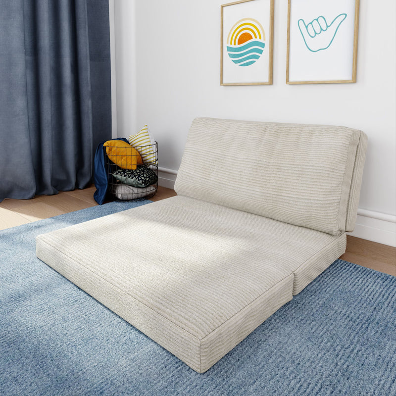 Beige ribbed fold-out floor sofa bed on a blue area rug in a modern minimal living room, wire basket of colorful throw pillows and framed wall art