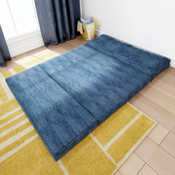 Blue foldable futon mattress on a wooden floor next to a yellow patterned rug, perfect for small spaces and modern living room decor.
