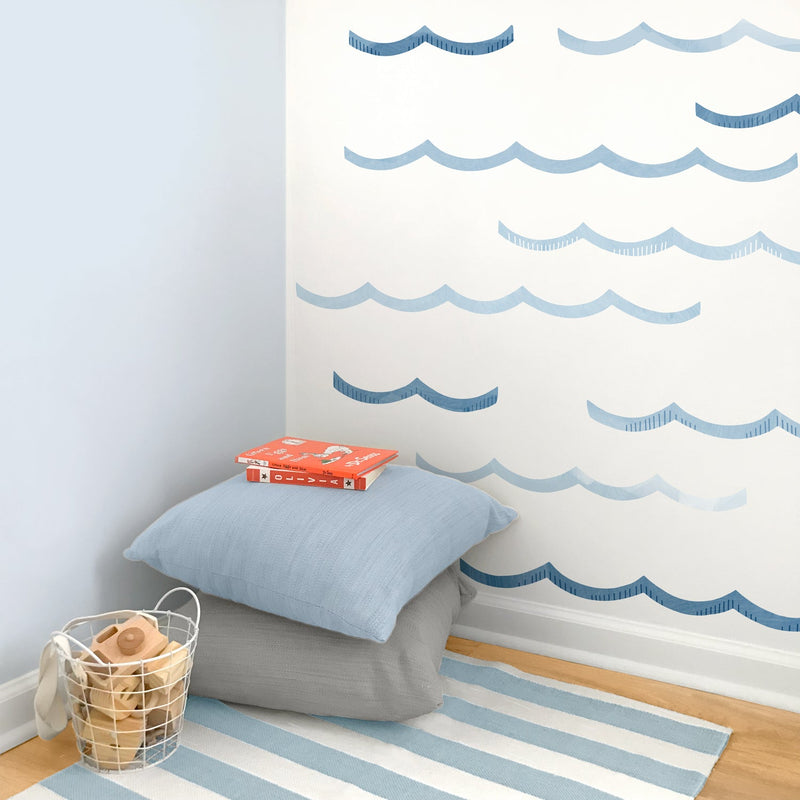 Coastal nursery corner with blue wave wall decals, striped rug, stacked pillows, wire basket of wooden toys and children’s books