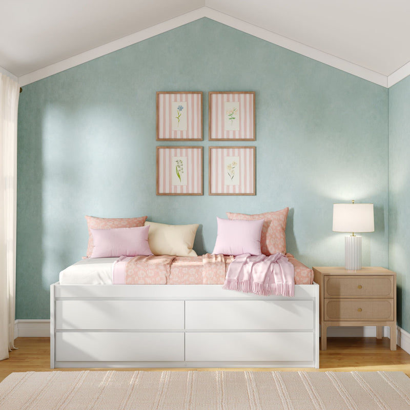 White storage daybed with pink floral bedding and pillows against a mint-green accent wall, framed botanical prints and a wooden nightstand with lamp, pastel bedroom decor