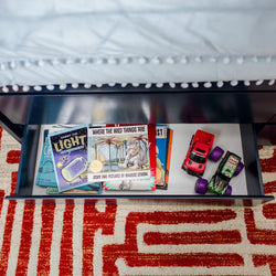 Open under-bed drawer in a kids' bedroom storing children's books including "Where the Wild Things Are" and two toy cars on a red geometric rug