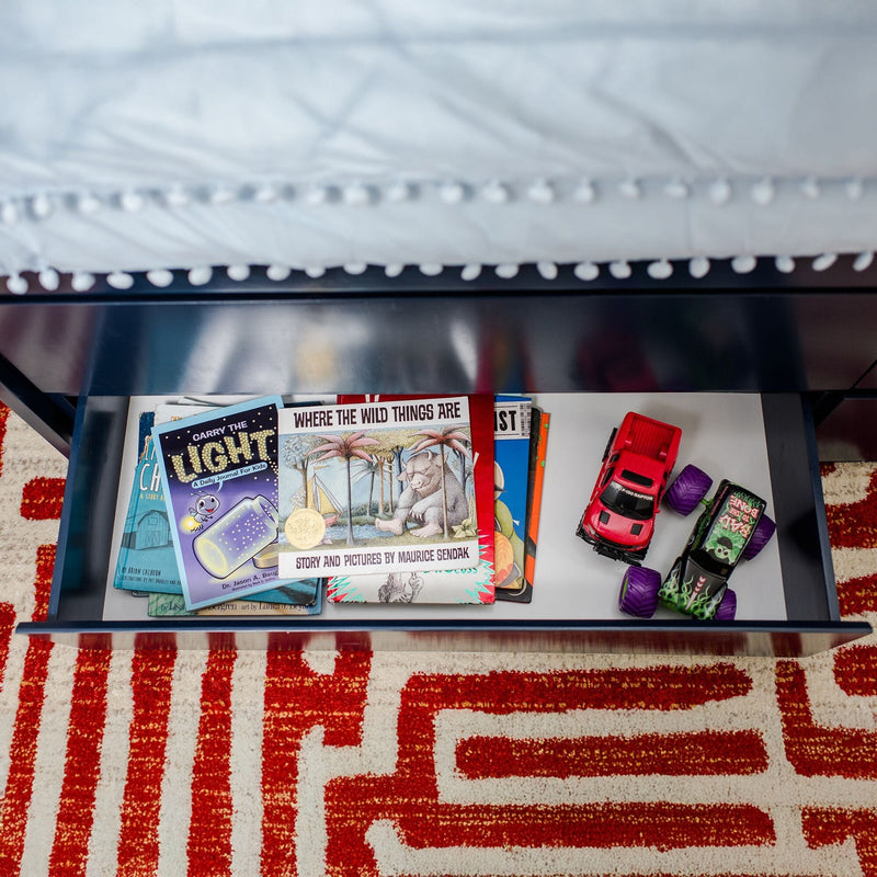 Open under-bed drawer in a kids' bedroom storing children's books including "Where the Wild Things Are" and two toy cars on a red geometric rug