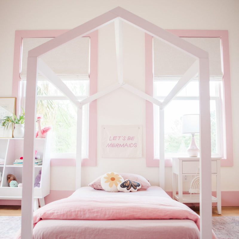 Children's bedroom with pink decor, featuring a white house-shaped bed frame, playful pillows, and a "Let's Be Mermaids" sign on the wall.