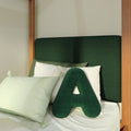 Close-up of modern bedroom with dark green textured upholstered headboard, wooden bed frame posts, green A-shaped accent pillow and pale green pillows