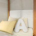 Neutral modern bedroom with textured linen headboard, pale yellow throw pillows, plush letter "A" cushion and wooden canopy bed frame