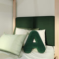 Close-up of green upholstered headboard with large green "A" pillow, light green decorative pillows and natural wood bed frame in a modern cozy bedroom