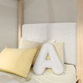 Close-up of a minimalist bedroom with a light-wood canopy bed, textured neutral upholstered headboard, yellow accent pillows and a white letter A decorative pillow