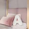 Blush pink upholstered headboard close-up with natural wood bedframe posts, soft pink gingham and textured mauve pillows, and a white decorative letter A cushion on pastel bedding