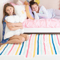 Two girls in a pastel kids' bedroom with pink bedding, decorative pillows, and a colorful striped rug — girls' room decor and cozy bedding inspiration