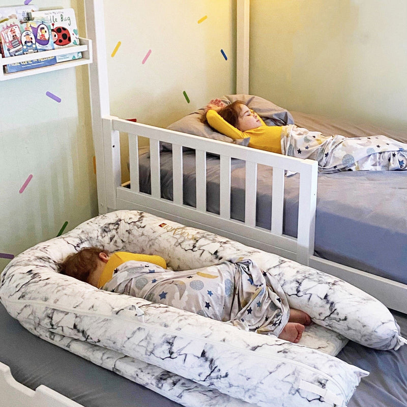 Two children sleeping in a cozy nursery featuring a white wooden toddler bed, marble-pattern baby lounger, star-and-moon swaddle blanket, gray bedding and colorful sprinkle wall decals