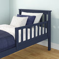 Navy blue wooden toddler bed with safety guardrail, navy quilt and white polka-dot sheets in a kids bedroom with light blue walls and hardwood floor