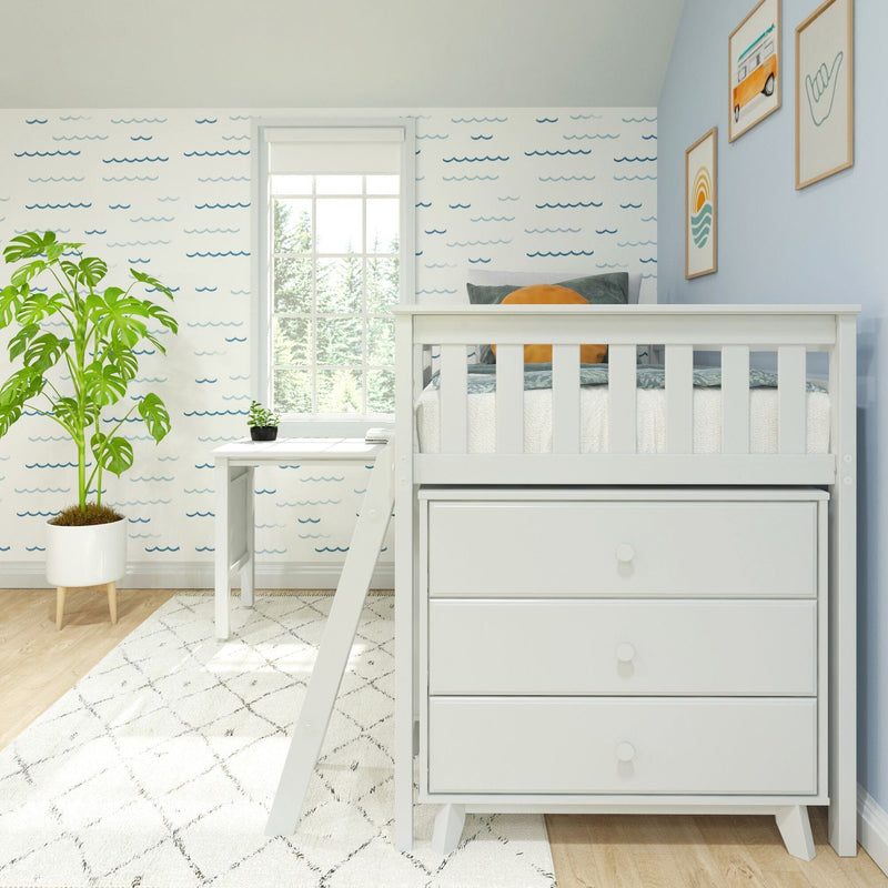 White kids loft bed with three storage drawers and ladder in a sunlit coastal-themed nursery, featuring blue wave wallpaper, white desk, large window, leafy potted plant and framed wall art