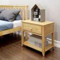 Light wooden nightstand bedside table with single drawer and lower shelf holding storage boxes beside a wooden bed with blue-gray bedding, topped by a gray house-shaped toy containing numbered blocks on a hardwood floor