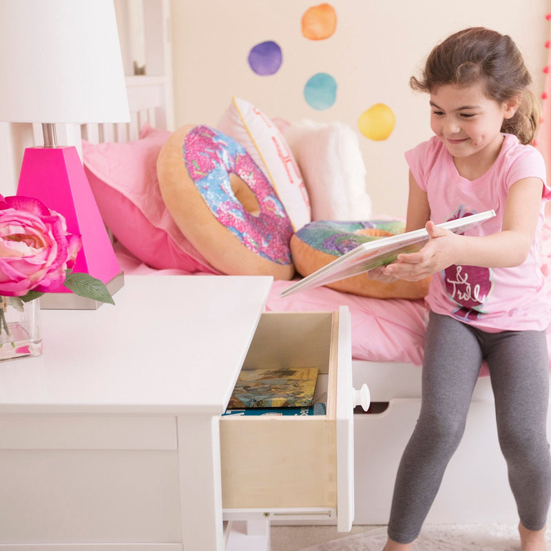 Young girl placing a book into a nightstand drawer in a pink kids bedroom with donut pillows and colorful wall decals — kids bedroom storage and organization idea