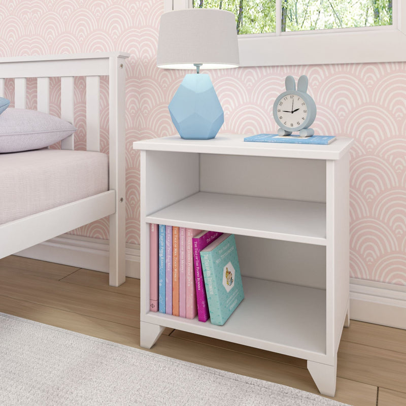 White kids' nightstand with books, blue lamp, and clock, beside a white bed in a pink-walled room.