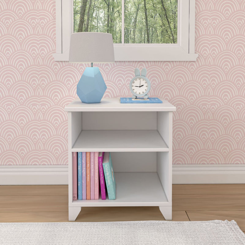 White nightstand with books, blue clock, and geometric lamp in a pink-patterned bedroom.
