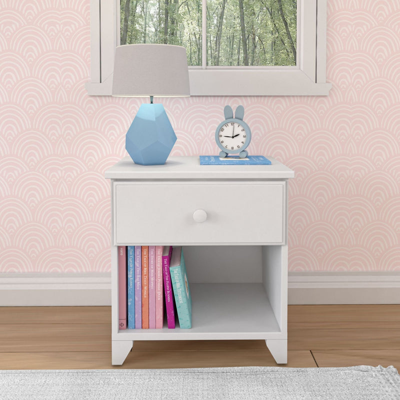 White nightstand with drawer, blue geometric lamp, bunny clock, and colorful children's books, set against pink wallpaper.