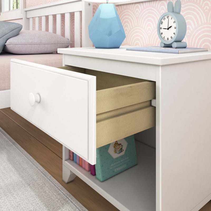 White bedside table with open drawer in a pastel kids bedroom featuring a blue geometric lamp, bunny alarm clock, pink patterned wallpaper and pillows, and children's books on the lower shelf