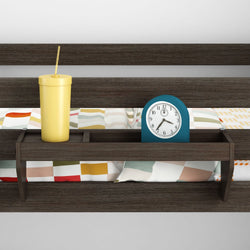Wooden bedside shelf attached to headboard holding a blue analog alarm clock and yellow tumbler with straw on a modern bed with colorful geometric bedding