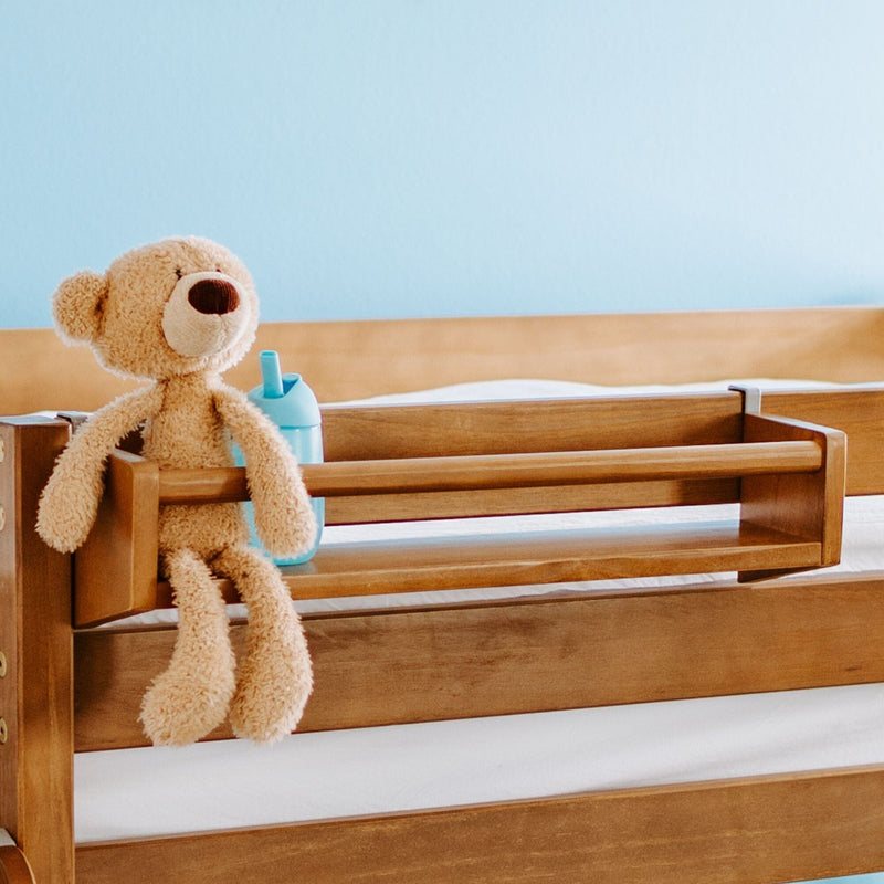 Wooden toddler bed with safety rail holding a plush teddy bear and blue sippy cup in a bright nursery