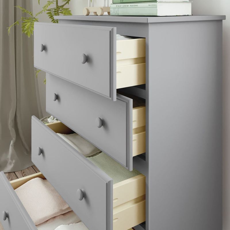 Gray wooden dresser with open drawers displaying neatly folded clothes, next to a plant and books for modern bedroom storage solutions.